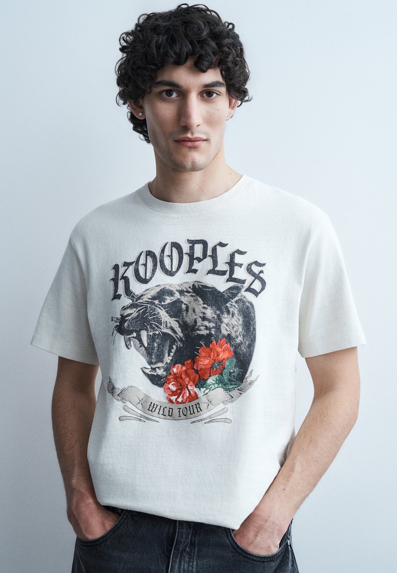 Νεαρός άνδρας με σγουρά μαλλιά που φοράει λευκό μπλουζάκι με σχεδιασμό λιονταριού που ουρλιάζει, κόκκινα λουλούδια και το κείμενο «KOOPLES WILD TOUR», με τα χέρια στις τσέπες.