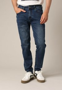 Homme portant un jean slim fit bleu foncé et des baskets blanches avec des rayures noires, se tenant avec une main dans la poche.