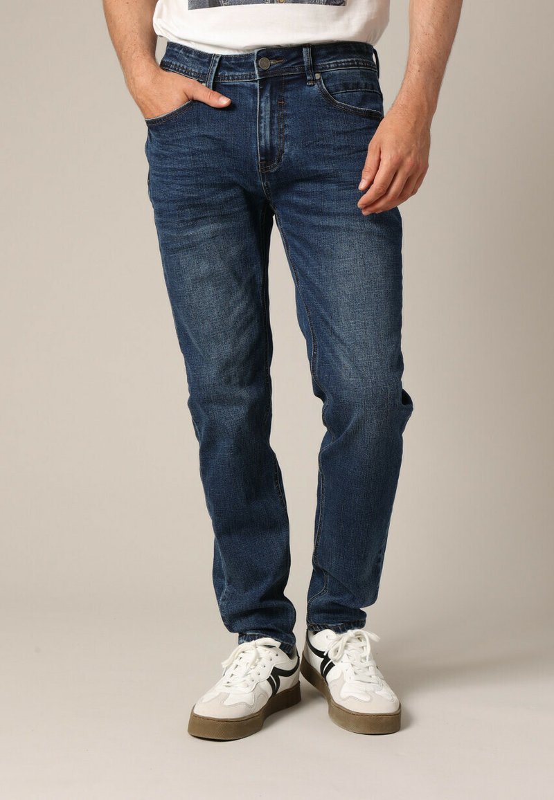 Homme portant un jean slim fit bleu foncé et des baskets blanches avec des rayures noires, se tenant avec une main dans la poche.