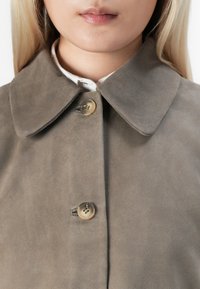 Detailní záběr ženy v béžovém semišovém kabátu s širokým límcem a dvěma průhlednými hnědými knoflíky zapnutými přes bílou košili.