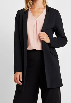 Vrouw draagt een lange zwarte blazer over een lichtroze V-hals blouse en zwarte wijde broek, houdt met één hand het blazerrever vast.