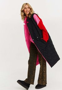 Czarna pikowana kamizelka z różowym podbiciem, zestawiona z czerwonym swetrem i spodniami w panterkę. Posiada długi fason, zatrzaski oraz boczne kieszenie.