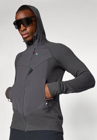 Hoodie zippé gris en tissu texturé. Dispose d'une capuche, de poches avant et de panneaux lisses contrastants. Les accents incluent un logo bleu.