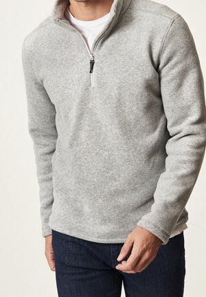 Hombre que lleva un forro polar gris claro con cremallera hasta la mitad, encima de una camisa blanca y vaqueros azul oscuro, de pie con la mano cerca del bolsillo.