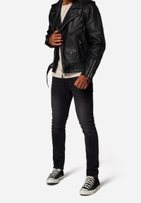 Černá kožená motorkářská bunda se stříbrnými zipy a kapsami, nošená přes světlé tričko, doplněná tmavými džínami a černými teniskami.