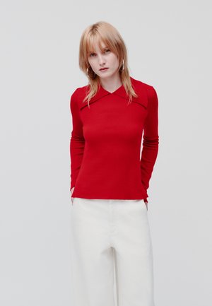 Blusa vermelha de manga longa com gola estruturada, design ajuste, detalhe em riscas nas mangas, combinada com calças largas em creme.