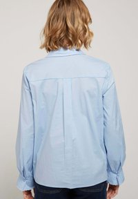 Chemise bleu clair avec col button-down, manches longues ornées de dentelle aux poignets, et un design de plastron à l'arrière.