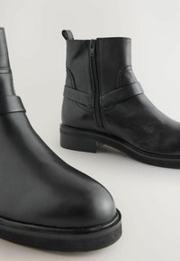 Botas de tobillo de cuero negro con una textura suave, punta redonda, cremallera lateral y detalle de correa decorativa. Suelas de goma con un tacón mínimo.