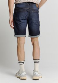 Donkerdenim shorts met opgerolde zomen, voorzien van een gladde textuur, achterzakken en een bruine leren label. Gecombineerd met witte gestreepte sokken en sneakers.