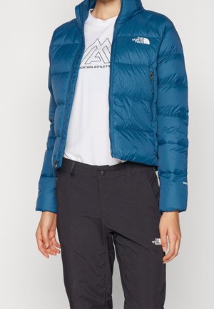 Personne portant une doudoune bleue, un t-shirt blanc Mountain Athletics et un pantalon noir North Face sur un fond uni.