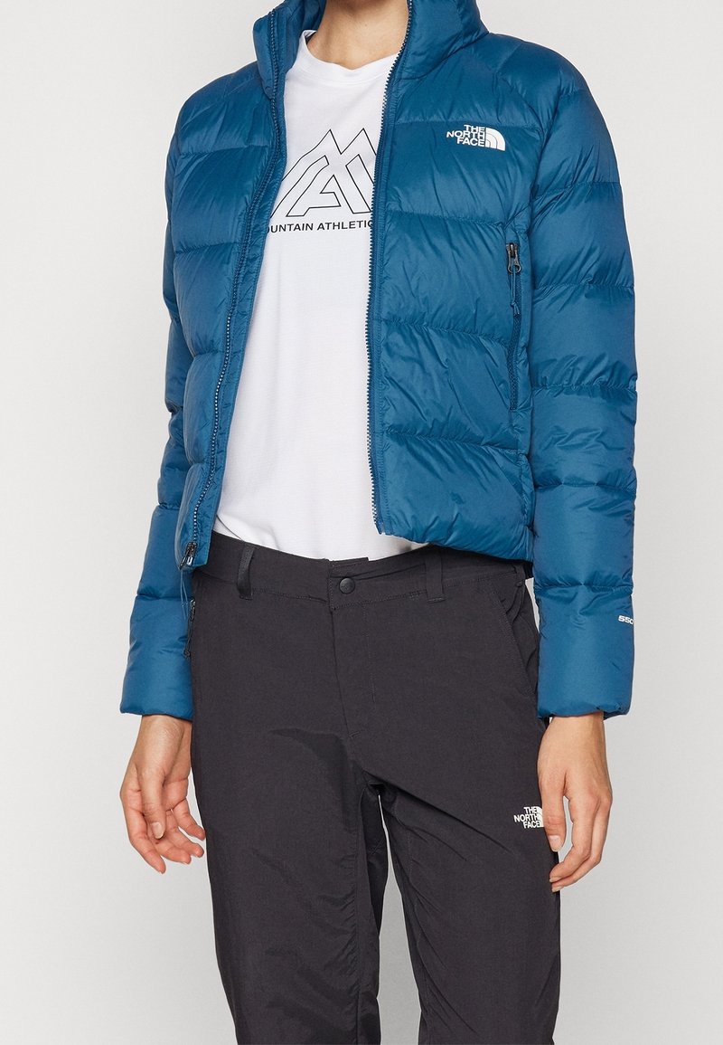 Personne portant une doudoune bleue, un t-shirt blanc Mountain Athletics et un pantalon noir North Face sur un fond uni.