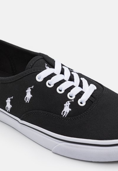 Sapatilhas de tela pretas com cordões brancos, logótipos da polo em branco bordados e uma sola de borracha branca com acentos pretos.