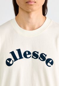 Camiseta de algodón color crema con logo "ellesse" en azul marino curvado en un estilo texturizado y bordado, con un clásico cuello redondo.