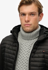 Junger Mann mit hellbraunem Haar, der einen grauen, strukturierten Rollkragenpullover und eine schwarze, gesteppten Jacke mit Superdry-Logo trägt.