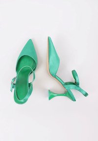 Zapatos de tacón alto de satén verde con punta afilada, correa en el tobillo y tacón de acrílico transparente, que exhiben una textura suave y herrajes decorativos.