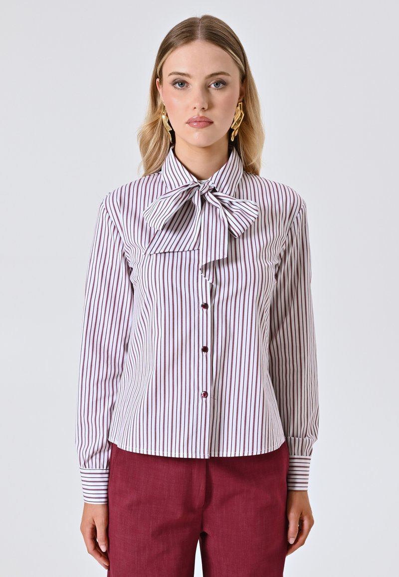 Blusa bianca con righe verticali bordeaux, caratterizzata da un ampio colletto a fiocco, chiusura con bottoni sul davanti e maniche lunghe.