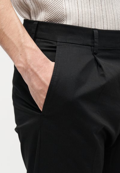 Main reposant dans la poche avant d'un pantalon noir sur mesure porté avec un pull en maille côtelée beige clair.