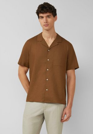 Młody mężczyzna z krótkimi kręconymi włosami, noszący brązową koszulę na krótki rękaw na guziki oraz jasnobeżowe spodnie na szarym tle.