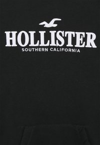 Svart hoodie i bomull, med vit broderad text "HOLLISTER SOUTHERN CALIFORNIA" och en liten måslogga ovanför texten.