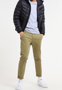 Svart puffjacka, grå skjorta, olivgröna chinos och vita sneakers. Jackan har en dragkedjeficka och ett kviltat mönster.