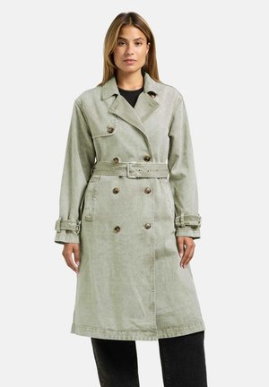 Kvinne iført en lys grønn dobbelspent trenchcoat med belte i midjen og mansjettstropper over svarte bukser, stående mot en ensfarget bakgrunn.