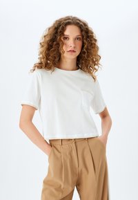 Camiseta blanca corta de algodón suave, con mangas cortas y un bolsillo en el pecho, con acabados cosidos a lo largo del dobladillo y los bordes de las mangas.