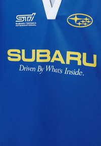 Tissu bleu avec logo Subaru, emblème STI et texte jaune « SUBARU Driven By What's Inside. » sous un col blanc en V.