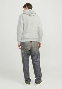 Grauer Kapuzenpullover mit geripptem Saum, kombiniert mit blauen Jeans mit einem ausgeblichenen Muster und weißen Turnschuhen vor einem neutralen Hintergrund.