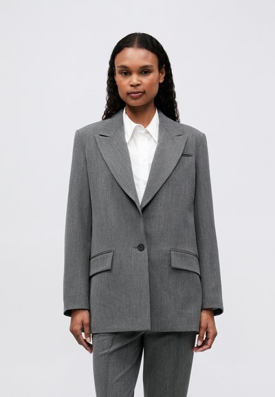 Blazer oversize gris en tissu texturé avec de grands revers, fermeture à un bouton et deux poches avant. Assorti à une chemise blanche.
