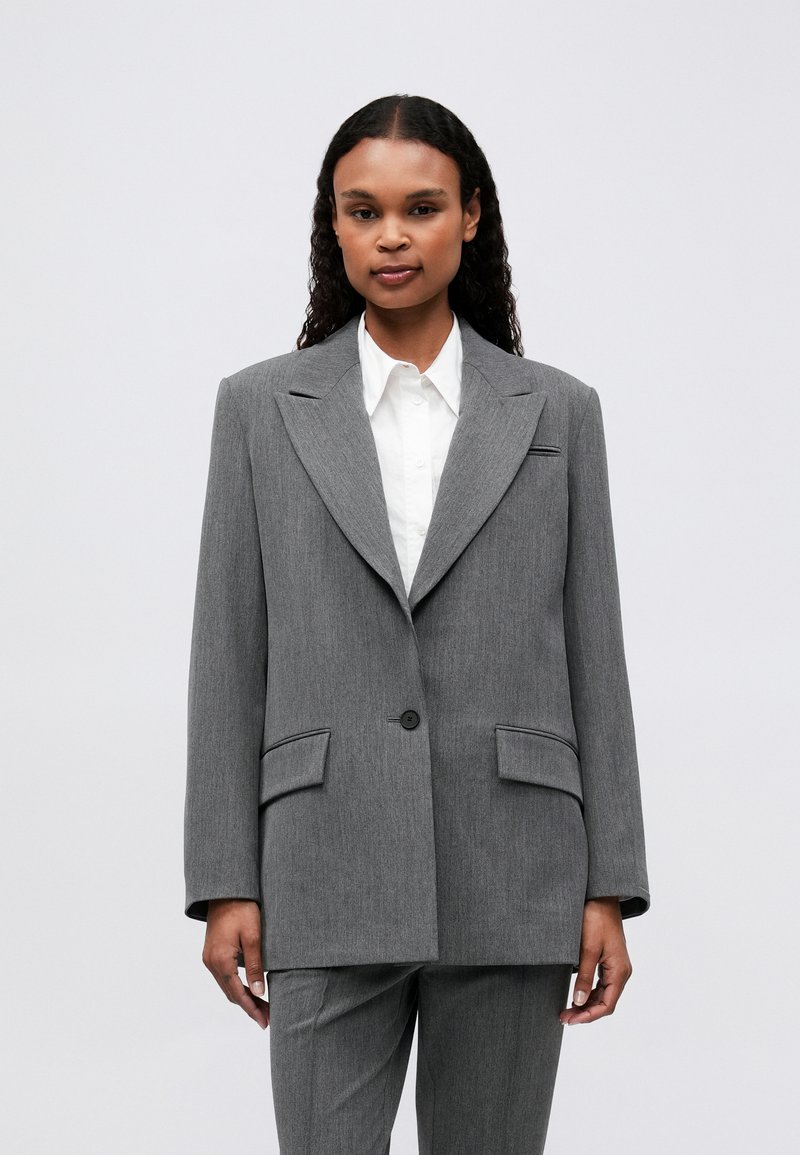 Blazer gri supradimensionat, realizat dintr-un material texturat, cu rever mari, închidere cu un singur buton și două buzunare frontale. Combinat cu o cămașă albă.