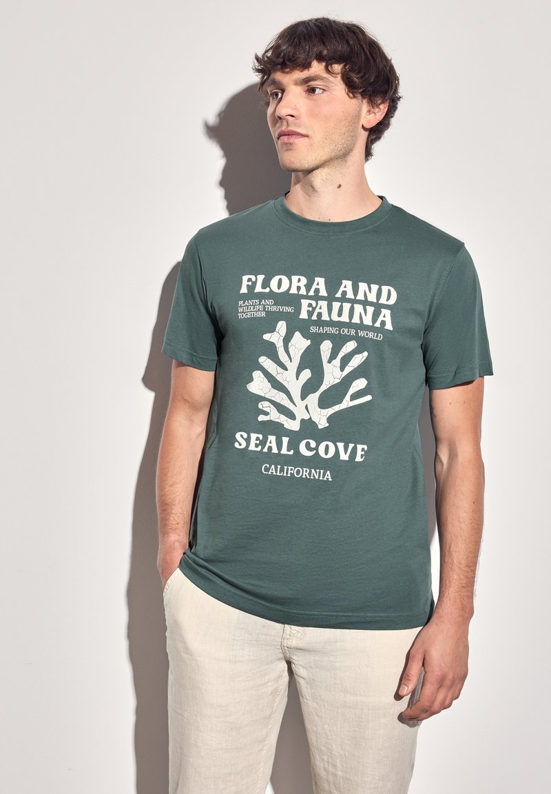 Zelené bavlněné tričko s bílým grafickým textem a korálovým designem. Navrženo s krátkými rukávy a kulatým výstřihem, kombinováno se světlými kalhotami.