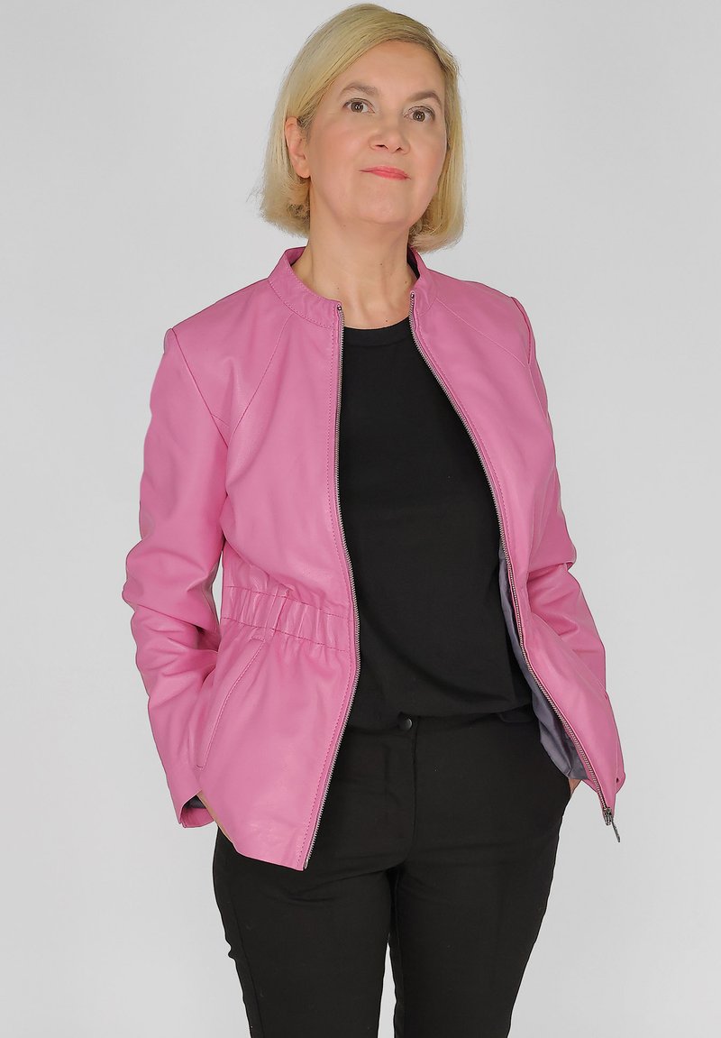 Rosa Lederjacke mit Reißverschluss, taillierter Passform und seitlichen Taschen. Über einem schwarzen Oberteil getragen, kombiniert mit schwarzen Hosen. Glatte Textur.