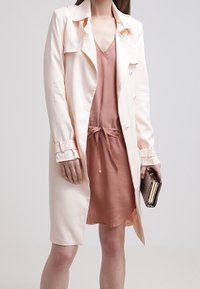 Długi, bladoróżowy płaszcz trencz z kołnierzem i zapięciem na guziki, noszony na bladoróżowej sukience z sznurkiem w talii. W ręku trzyma teksturowaną torebkę.