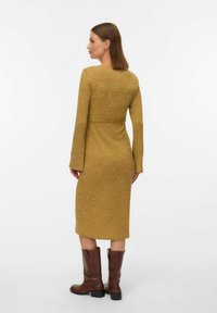 Dzeltenīgs ribots adījuma kleita ar garām piedurknēm un pieguļošu siluetu, savienota ar ceļa augstumā brūnām ādas zābakiem.