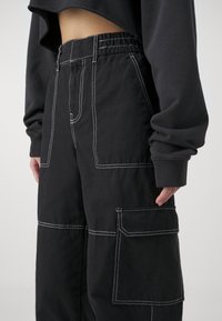 Persoon in zwarte cargobroek met witte stiksels en een zwart langemouwige cropped top, hand rustend bij de zak.