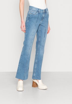 Femme portant un jean droit bleu clair avec des détails de couture à l'avant et des mocassins blancs à talons, debout devant un fond clair uni.