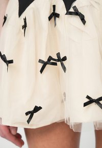 Kreemi tülli kleit mustade satiinivöödega. Omab kihilist tekstuuri ja laienevat vormi. Seeliku äär on kergelt läbipaistev.