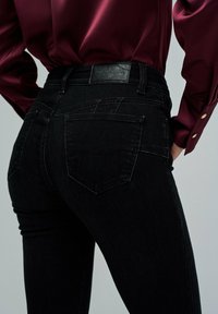 Acercamiento de una persona que lleva puestos unos pantalones vaqueros negros de talle alto con bolsillos traseros y una camisa de manga larga burdeos metida por dentro.