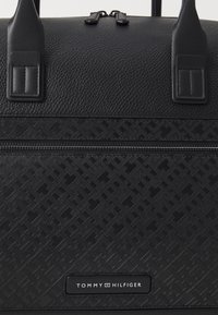 Sac de voyage noir texturé avec deux poignées supérieures, comprenant un compartiment à fermeture éclair et un motif géométrique en relief. Logo sur le panneau avant.