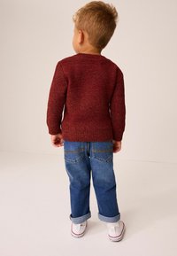 Stickad vinröd tröja med en texturerad yta, ihopparad med uppvikta blå jeans som har gula sömmar på bakfickorna och vita sneakers.