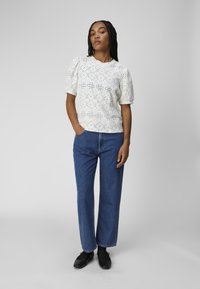 Witte blouse met korte mouwen en een oogjespatroon, gecombineerd met hoge taille rechte spijkerbroek en zwarte platte schoenen.