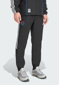 Schwarze Sporthose mit weißen Streifen, elastischen Bündchen und einem bunten gestickten Logo auf dem Oberschenkel. Tragen mit grau-weißen Turnschuhen.