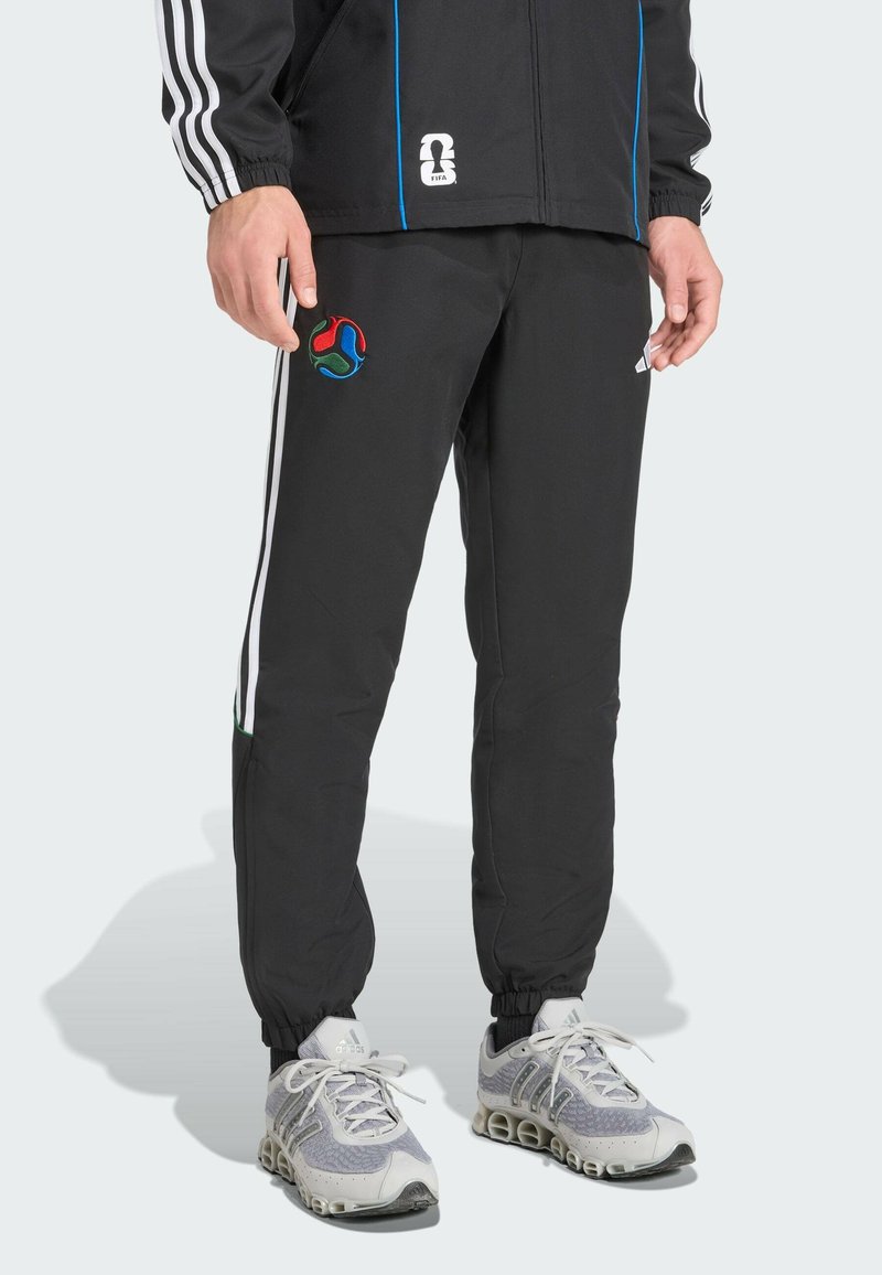 Schwarze Sporthose mit weißen Streifen, elastischen Bündchen und einem bunten gestickten Logo auf dem Oberschenkel. Tragen mit grau-weißen Turnschuhen.