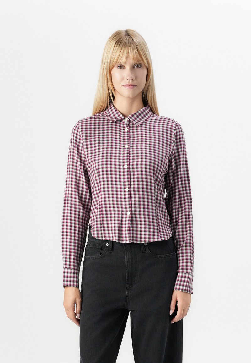 Button-up shirt in checkered pattern with purple and white squares, made of cotton. Features a collar and long sleeves paired with dark jeans.