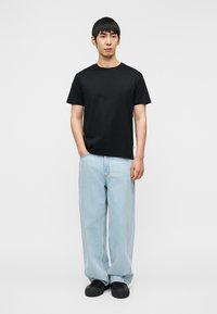 T-shirt en coton noir associé à un jean bleu clair à coupe ample. Le modèle porte des baskets noires, debout devant un fond blanc uni.