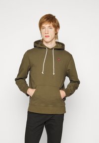 Felpa verde oliva con cappuccio regolabile, tasca frontale a marsupio e un piccolo logo rosso. Maniche lunghe con polsini a costine, realizzata in tessuto morbido.