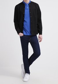 Camisa negra de botones sobre un polo azul, combinada con jeans oscuros y zapatillas blancas. El conjunto presenta un diseño casual con una forma ajustada.