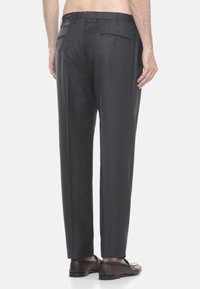 Uomo che indossa pantaloni eleganti grigi scuri e mocassini in pelle marrone, visto da dietro su uno sfondo bianco.