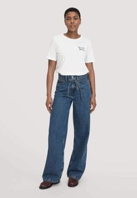 Witte katoenen t-shirt met korte mouwen en een kleine opdruk, gecombineerd met hoge taille wijdvallende blauwe spijkerbroek en bruine schoenen.