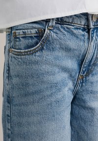 Primer plano de unos jeans de mezclilla azul claro con costuras detalladas y bolsillo lateral, combinados con una blusa blanca.